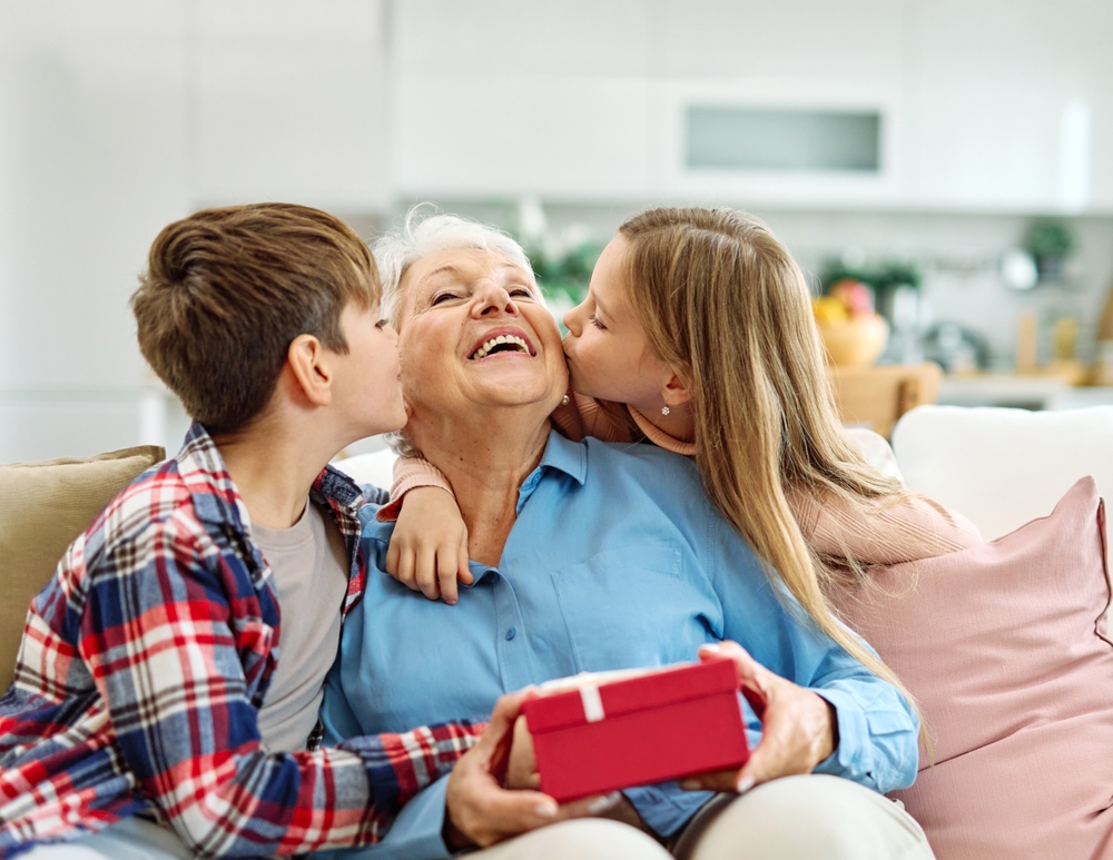 Granddaughter,And,Grandson,Surprises,Her,Grandmother,With,Present,On,Mother's, Couch, Gift