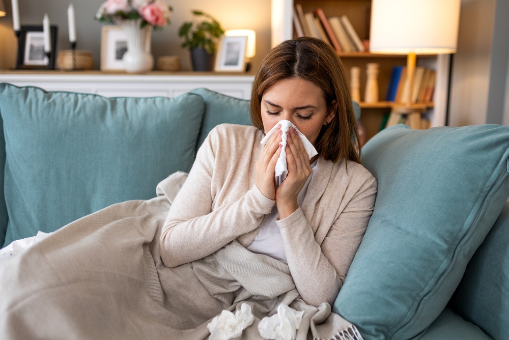 Woman sneezing into a kleenex tissue on teal couch.